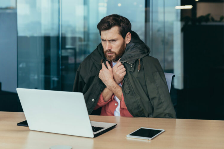 It's cold in the office, the businessman is freezing while working on a laptop inside the building, the man is covered with a warm jacket.